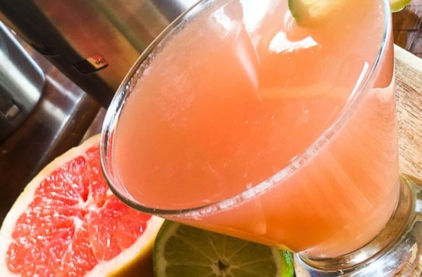 A pink grapefruit martini with ice crystals on the glass, garnished with a grapefruit slice and mint leaves.